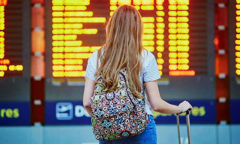 The back of someone with a backpack on looking at an airport arrivals and departures board.