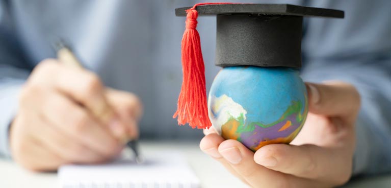 someone's writing on paper with one hand and the other hand holding a small globe, with a graduation hat on top