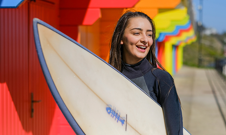 Female in a wetsuit holding a surfing board in front of beach huts