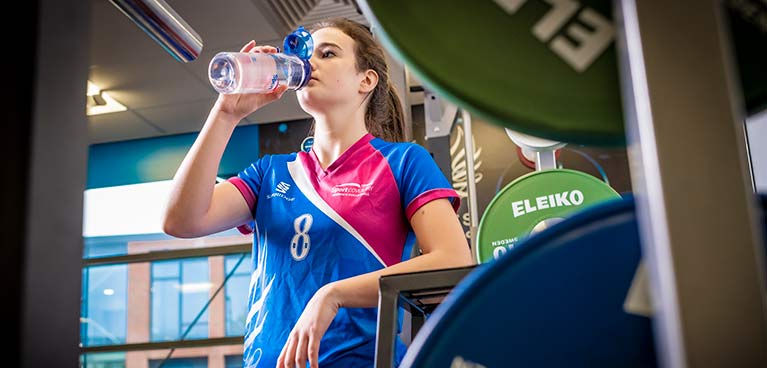 A female student in a sports team top, drinking from a bottle of water.