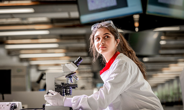 A student in a science lab, wearing a white lab coat and gloves with safety goggles on top of her head, looking up from a microscope.