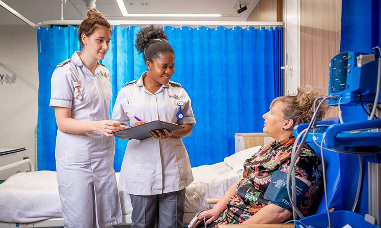 Two student nurses talking to a mock patient in a mock hospital bay