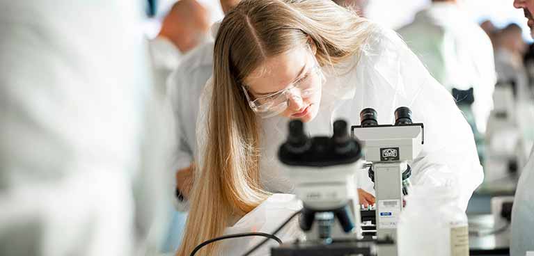 Student in a lab coat and goggles looking into a microscope