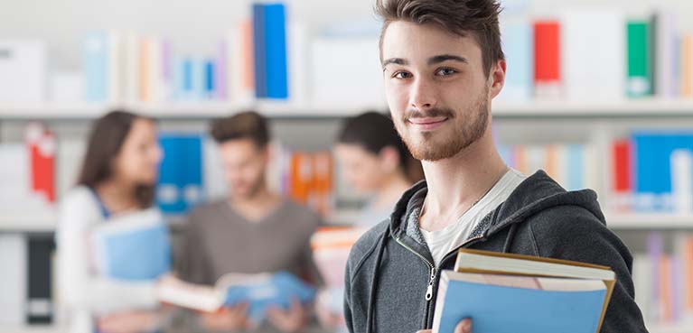 A student holding books smiles, three blurred students talking behind him