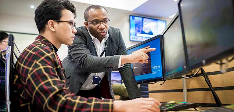 Student in the Trading Floor A student using a computer at a desk wearing glasses next to a tutor pointing at the screen