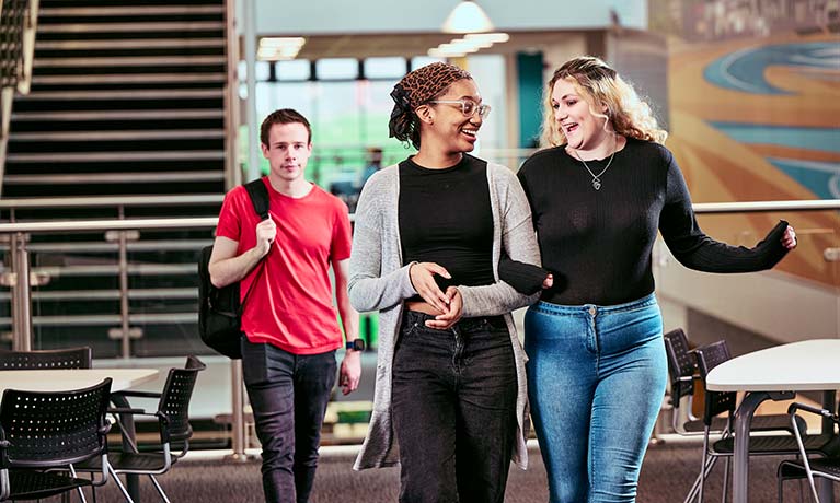 Two students walking together linking arms with another student walking behind