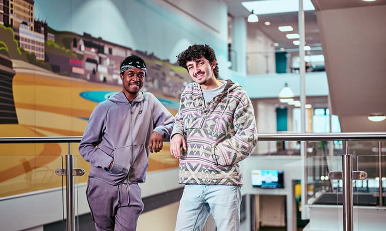 Two male students leaning against a balcony looking at their phone
